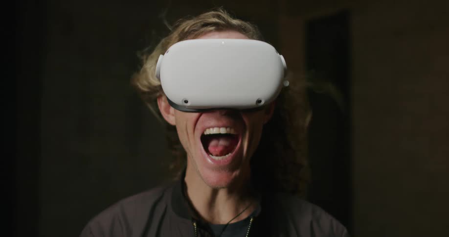 A man with long blond hair wearing a virtual reality headset screams as his hair is blown backwards