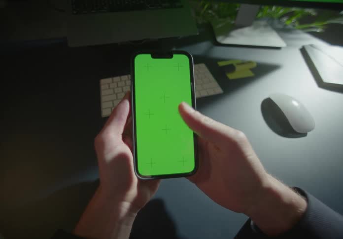 Man with black shirt holds smartphone with green solid screen sitting at desk