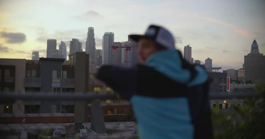 A man looks out over a cityscape while leaning against a chain link fence on a rooftop