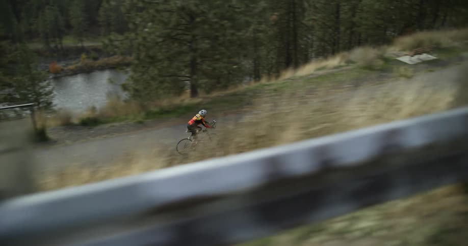 A bike rider passes another on a path at the bottom of a grassy slope with a river behind