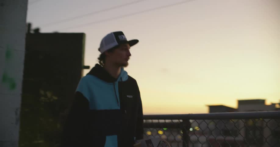 A man carries a skateboard as he exits onto the rooftop of a building in a city at sunset