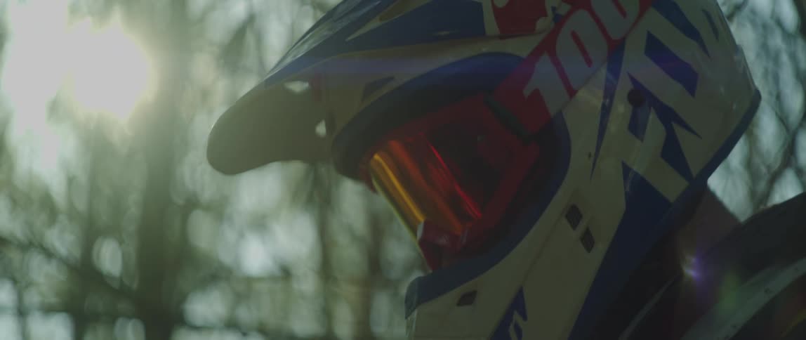 A person wearing a crash helmet and goggles is outside as the sun shines through the trees