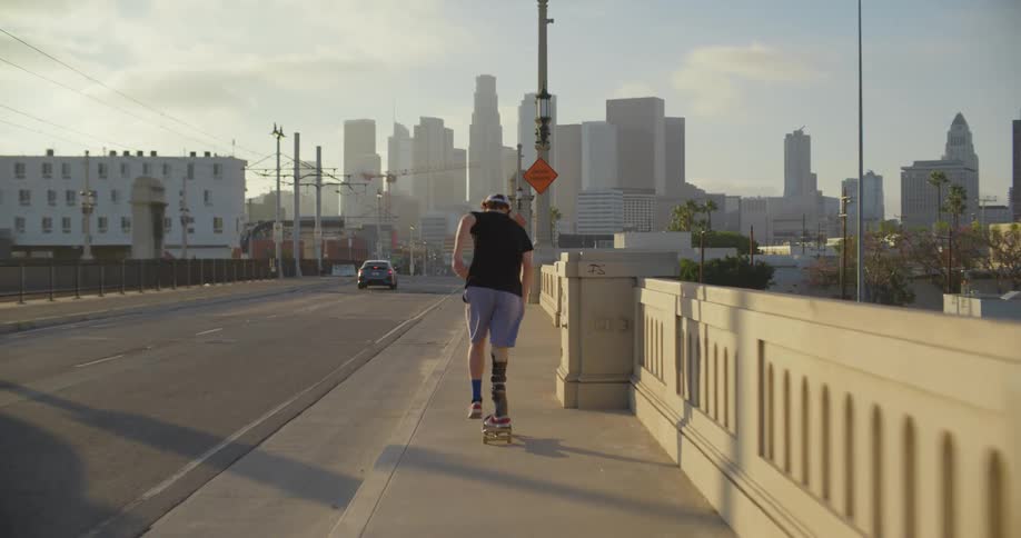 A young male skateboards on a bridge sidewalk toward a large city skyline