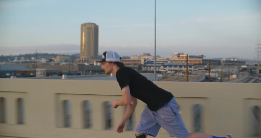 A man flips his skateboard while skateboarding across a bridge over a train track in a city during the day