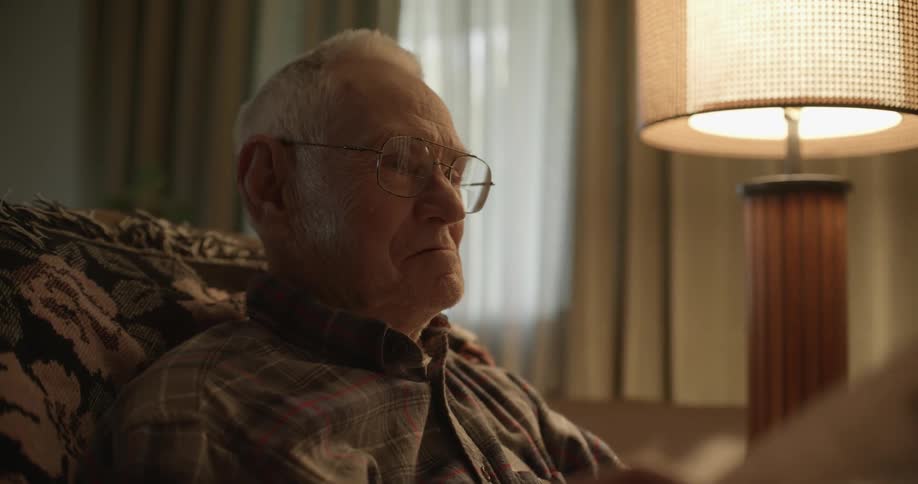 Elderly man sitting on a recliner reading the newspaper next to a lamp