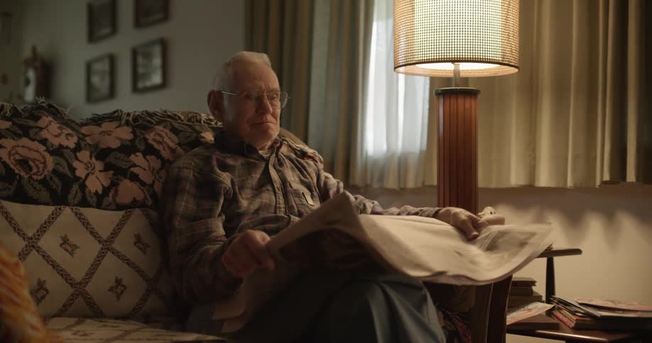 Elderly man with glasses and plaid shirt and trousers reads newspaper and looks at his granddaughter in living room at home