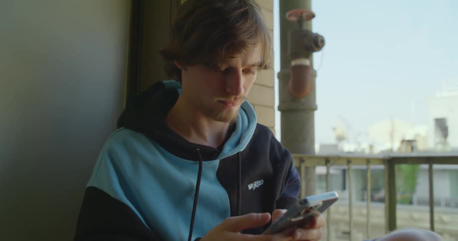 A man is sitting on a balcony texting on his cell phone
