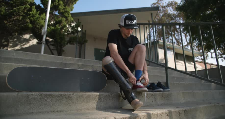 A teenager with a prosthetic leg puts on a shoe as he sits on the steps of a building