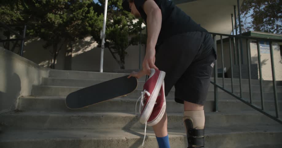 A man with a prosthetic leg carries a skateboard and a pair of shoes and sits down on some steps outside