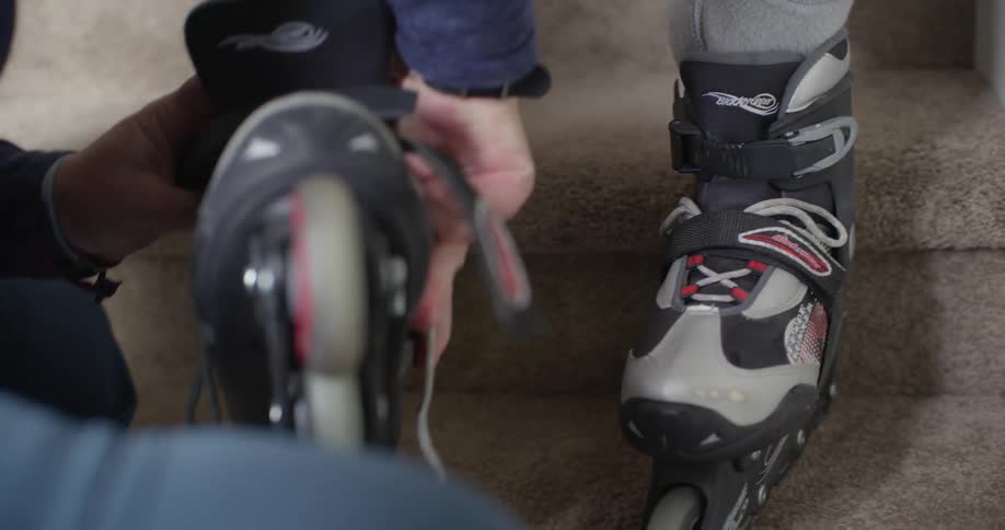 An adult is helping a child put on roller skates as they sit on some carpeted stairs