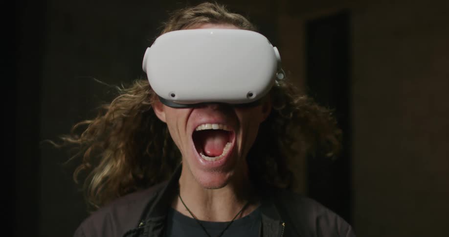 A man with long blond hair wearing a virtual reality headset screams as his hair is blown backwards