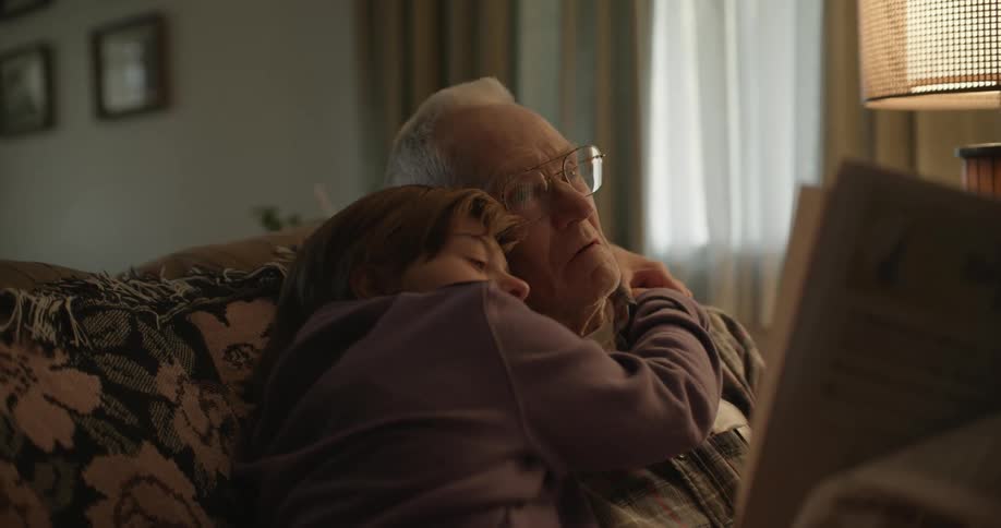 A girl leans in to hug his grandfather as they read a newspaper on the couch