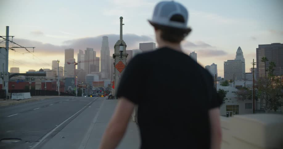 A teenage boy skateboards down a city sidewalk towards the downtown skyline