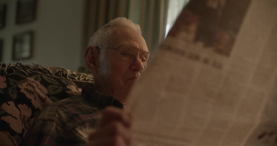 Elderly sad man with glasses and dark shirt sits on sofa reading newspaper