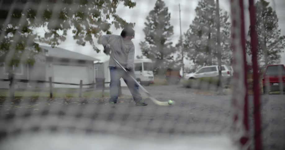 A kid is on a neighborhood sidewalk hitting a ball into a net with a hockey stick