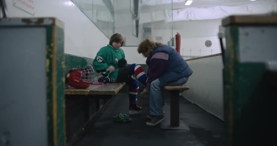A woman sitting on a bench ties her sons skates for him