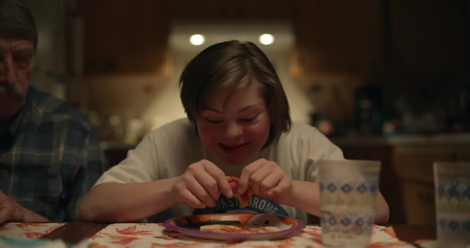 A girl sits at the dinner table with her family and laughs as her pizza falls on her face