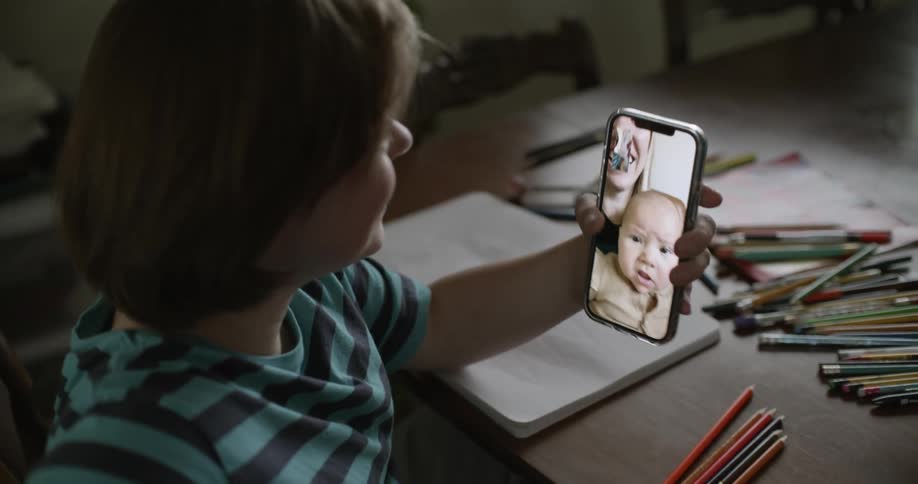 Girl with striped shirt sitting at table makes video call on smartphone with mother and baby in room