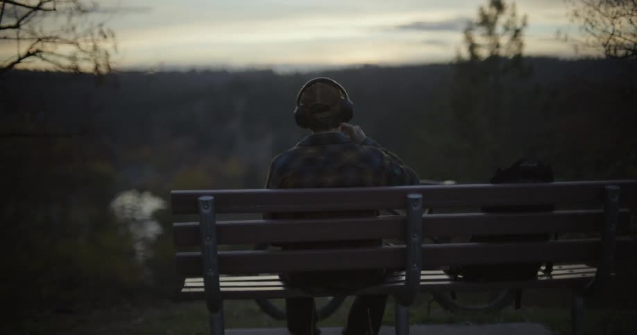 Young man sitting on a bench over a hill looking at the sunset while drinking from a bottle