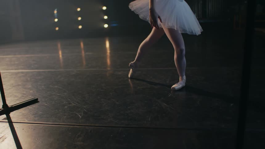 A ballerina is dancing a ballet on a dark stage under lights in an empty theater
