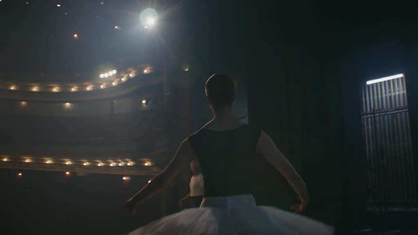 Young ballerina enters the stage of an empty teather holding a pose and standing in front of a trainer