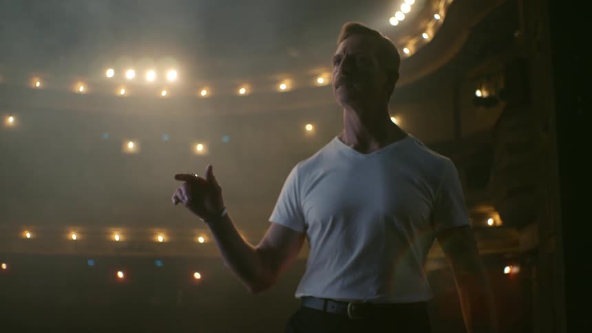 A man stands on a stage and looks straight ahead as he moves his arm up and down