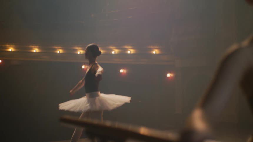A ballerina dances across a stage while another one stands at a barre and practices poses