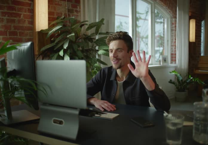 A man is sitting at a desk with a computer waving to someone in a video conference