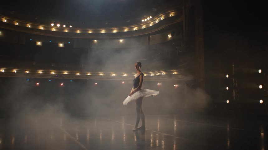 A ballerina posing on a misty stage in theater turns to look into the camera
