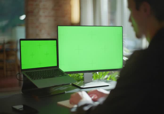 A man sitting at a desk is typing on a keyboard in front of a monitor and a laptop