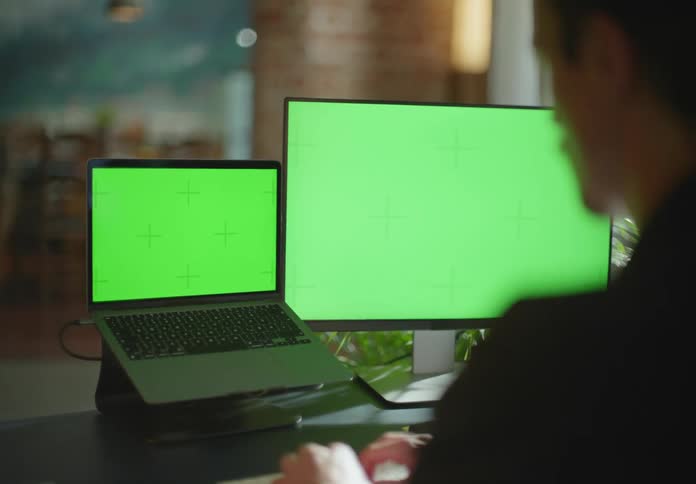 Young man typing on the keyboard of a laptop with an extra monitor with green replaceable screens