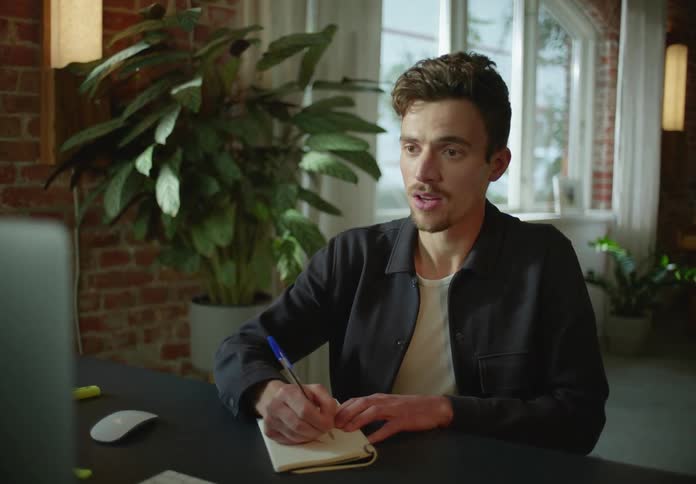 Man with goatee black jacket and white shirt looks at laptop smiling and writing down in notenook