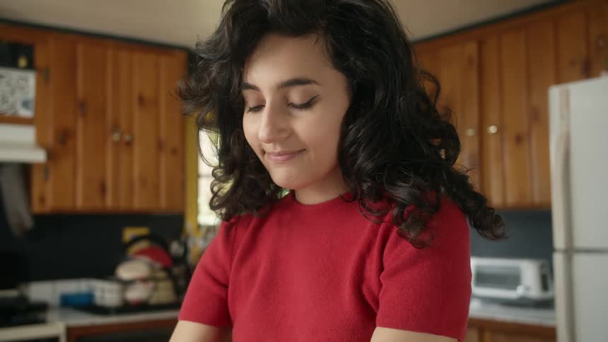 Happy woman with black wavy hair and red shirt looks down making sandwhich in kitchen