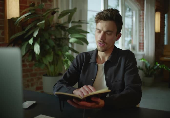 A man sits in front of a computer at home and writes in a book