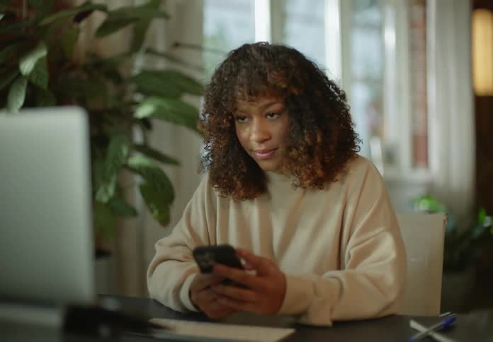 Woman with brown curly hair and beige turtleneck sweater looks at laptop and uses smartphone next to big houseplant in room