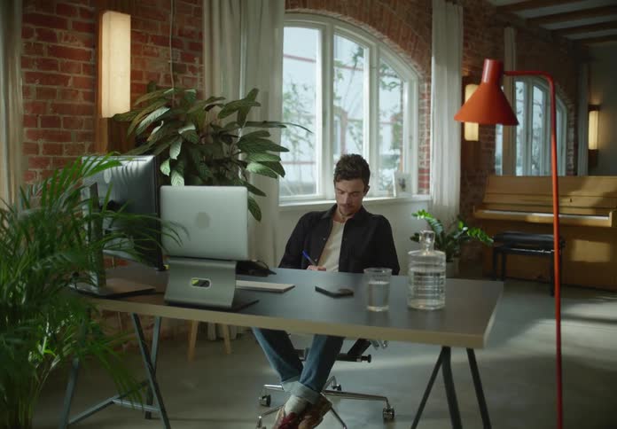 Man with black jacket white t shirt and jeans sitting at desk looks at laptop and writes in notebook