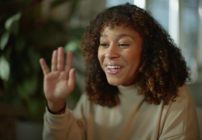 Woman with brown curly hair and beige turtleneck sweater smiles and waves at person on video call in room