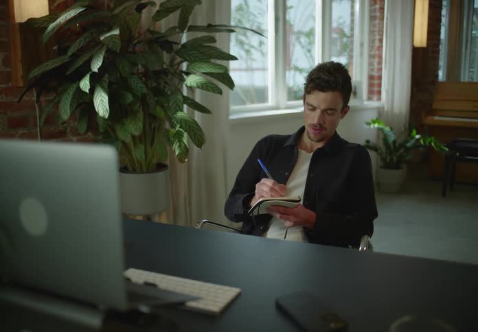 Man with goatee black jacket and white shirt writes down in notebook looking at laptop next to brick wall and houseplant in room