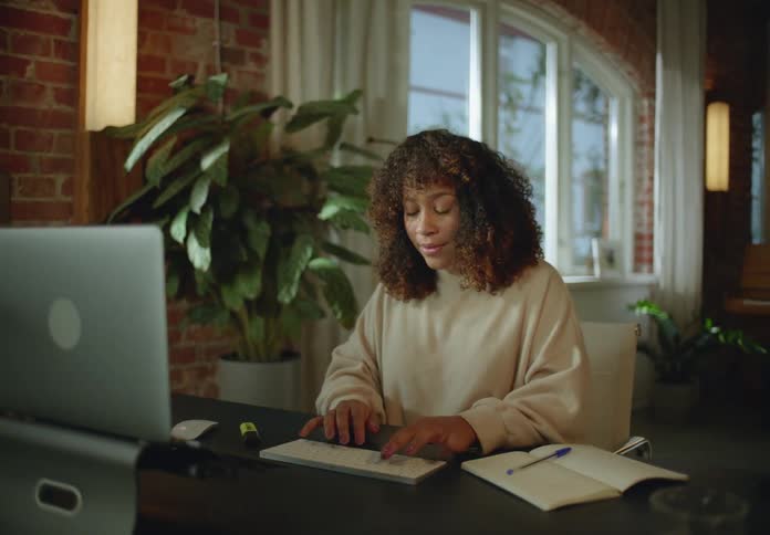 Woman types on keyboard at desk with piano and houseplants in the background