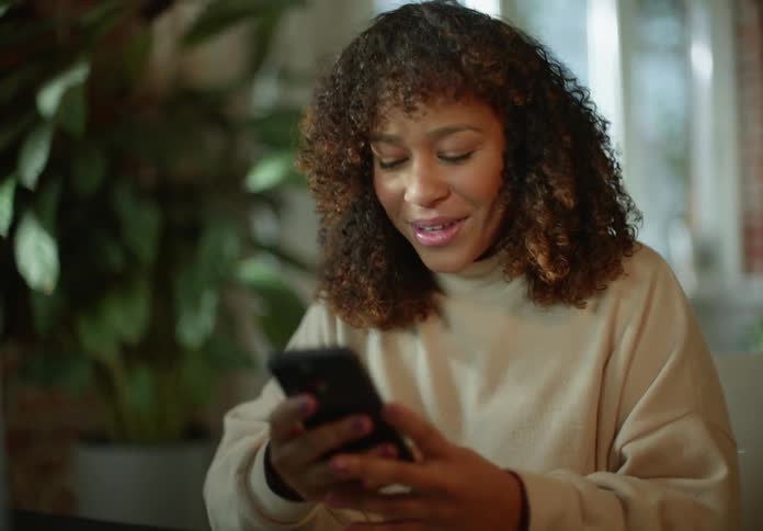 Woman with curly hair and beige turtleneck sweater talks on video call on laptop and texting on phone in room