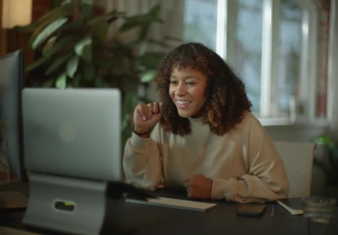 Woman with brown curly hair and beige turtleneck sweater waves at person on video call on laptop in room
