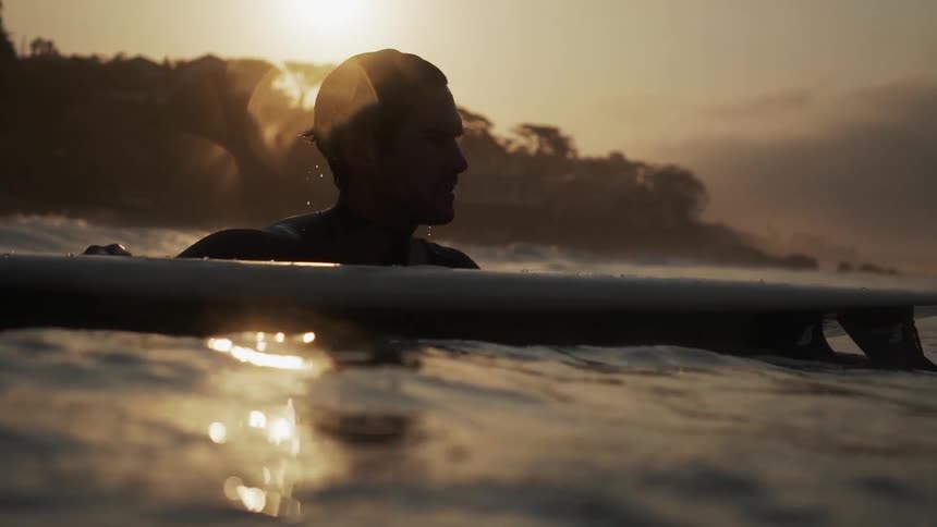 A man holds on to a surfboard near sunset