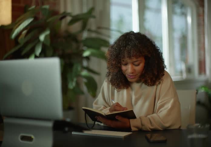 A woman is sitting at a desk in front of a computer having a video conference and writing in a book