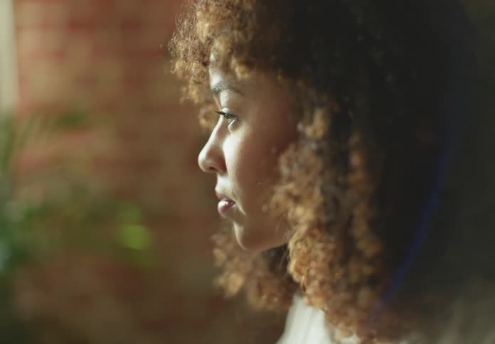 Woman with brown curly hair and beige turtleneck sweater works on laptop next to brick wall with houseplant