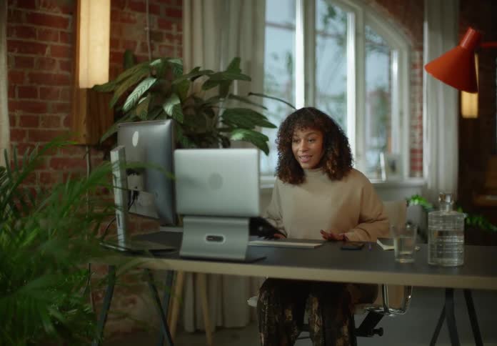 Woman with curly hair and beige sweater sits at desk making a video call on laptop next to brick wall in room