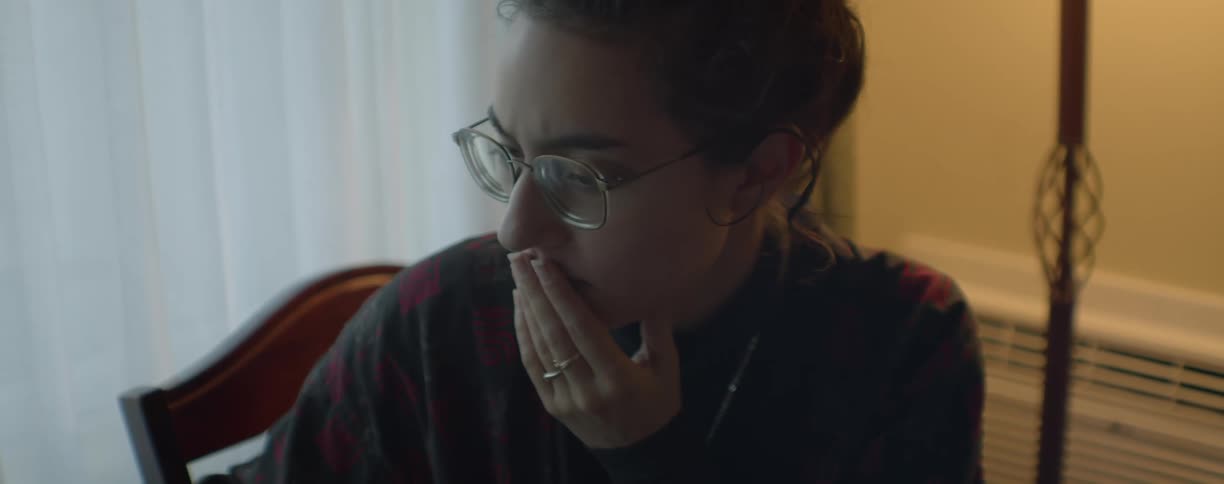 Woman with glasses and black blouse sits in chair putting her hand on her mouth next to curtains in room