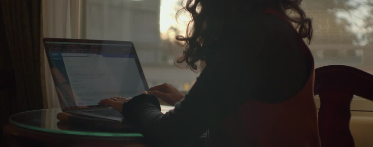 Woman with black wavy hair shirt and brown vest sits at table working on laptop in room