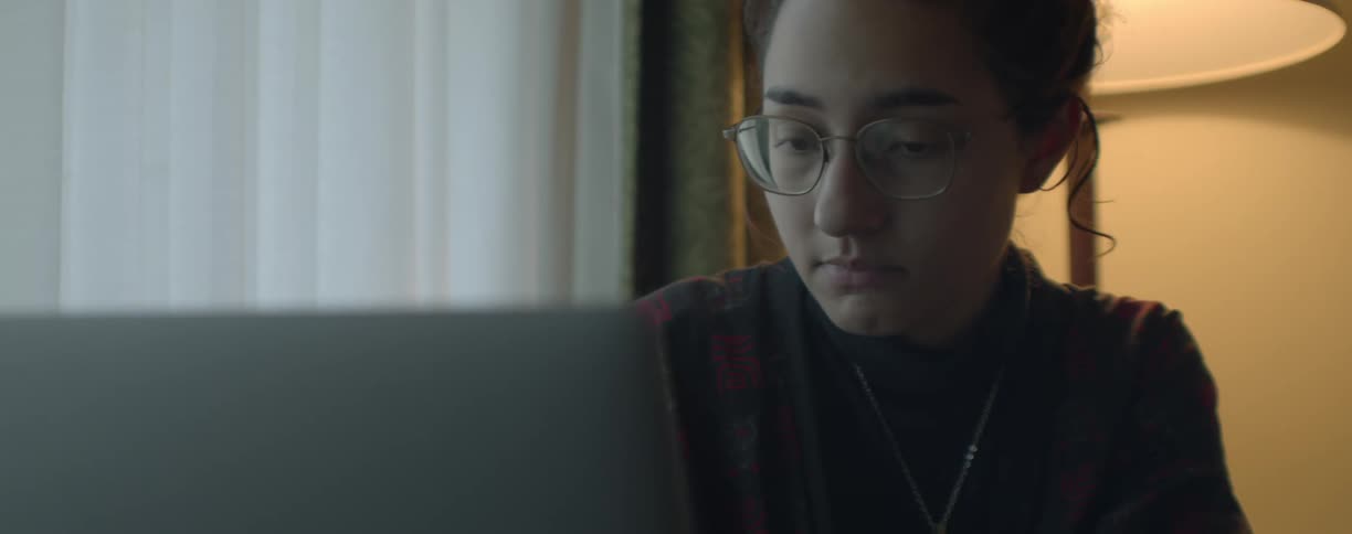 Woman with glasses and black blosuse sits at desk working on laptop next to white curtains
