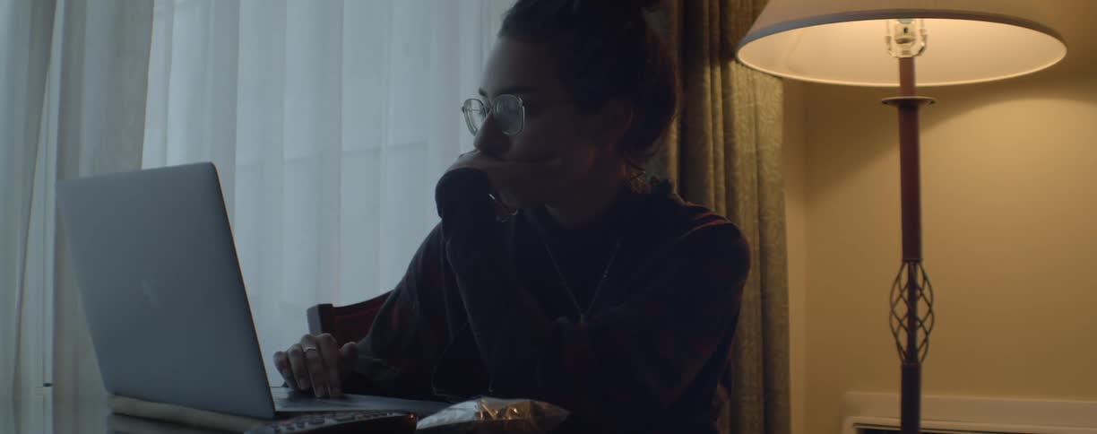 Woman with her hair in bun and glasses works on laptop next to white curtains