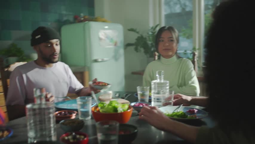 A group of people chat while eating together at a table in a kitchen during the day